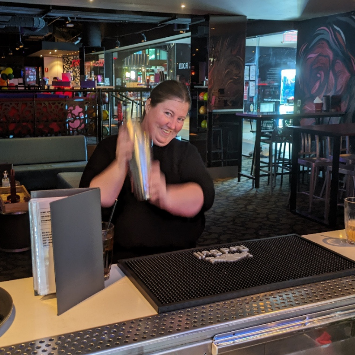 A woman smiles as she shakes a cocktail mixer over a bar.